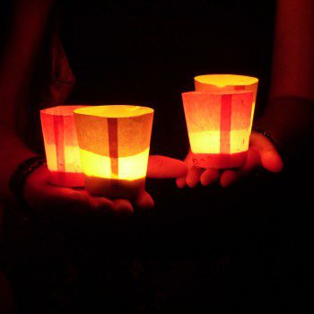 Outstretched hands hold small paper bags illuminated by candles.