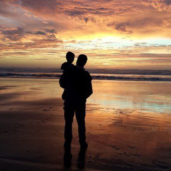 A man holds a young child while standing on a beach looking at a setting sun.