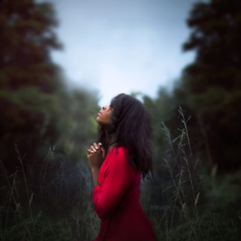 A woman in a red dress looks up towards the sky in prayer.