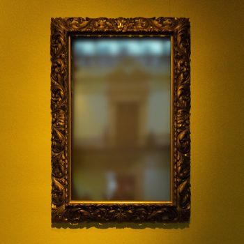 A blurry image in a mirror with a gold frame, hanging on a yellow wall