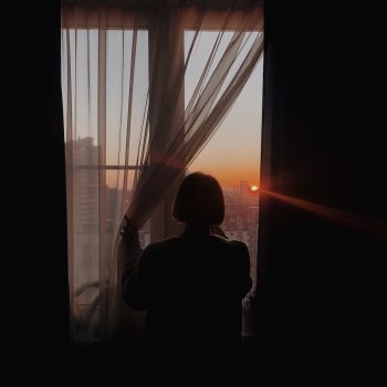 A woman holds back a curtain and peers out a window at sunset