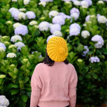 Person with dark hair and a yellow knitted cap and pink sweater faces a large bush of purple hydrangeas.