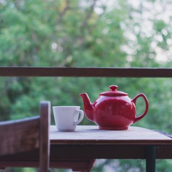 A bright red teapot and white cup sit on a wooden table outside on a porch.