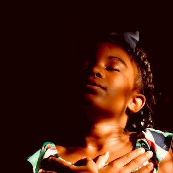 Young person with dark, braided hair, and eyes closed feels the sun's warmth and glow on their face.