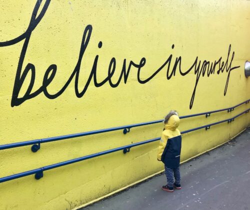 A child in a winter coat facing a yellow wall that says "believe in yourself" in script lettering