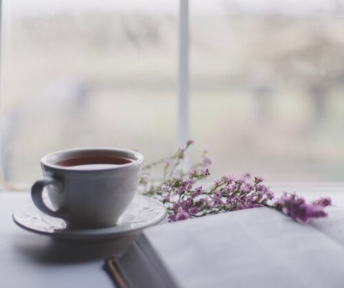 A white ceramic mug of coffee on a table near a book and small purple flowers
