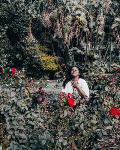 Woman sitting in a garden of red roses with her eyes closed, hand on her heart, and a smile on her face