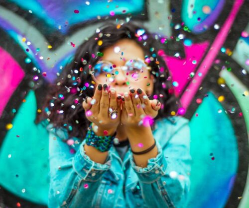 A woman in a denim jacket blowing colorful confetti out of her hands