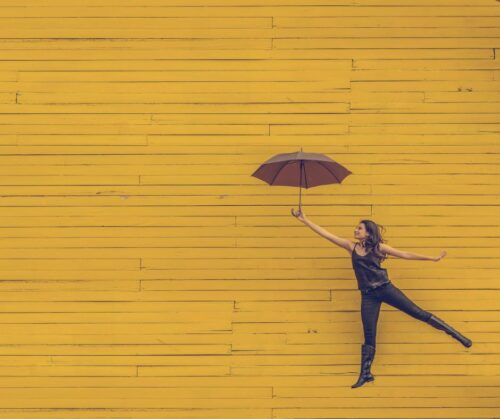 A woman holding an open umbrella jumping elegantly in front of a yellow wall