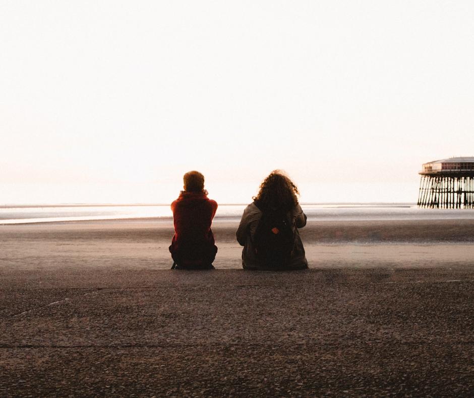Two people sitting on a beach wearing coats while away from the camera