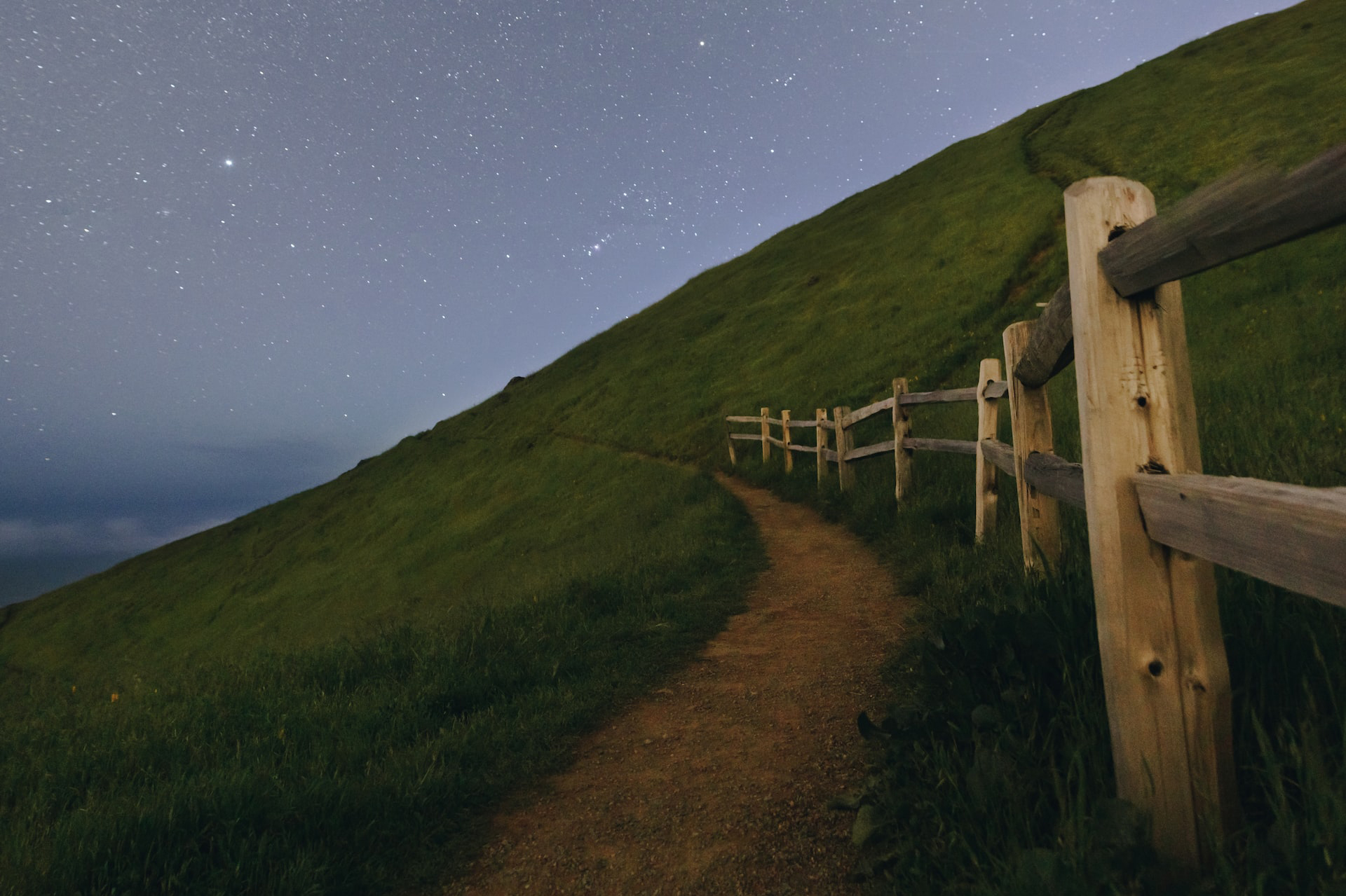 Wooden fence along a pathway on a hillside at nighttime with stars in the sky