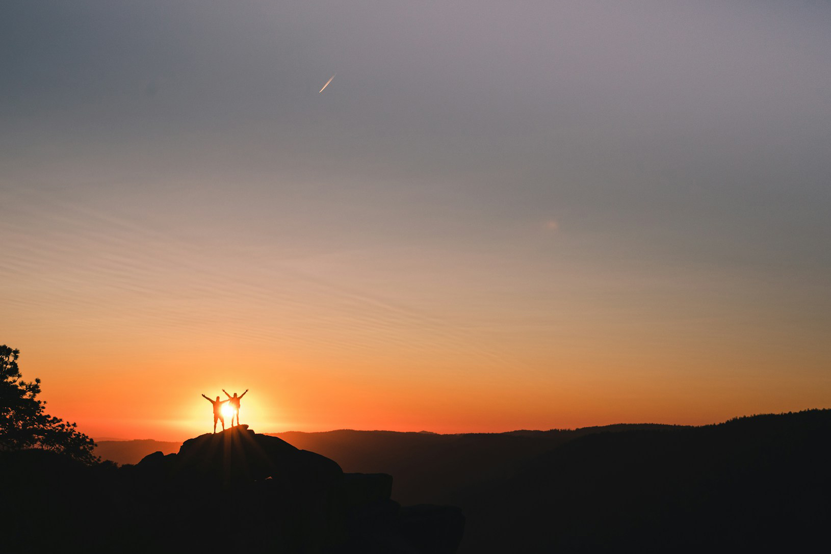 Silhouette of a couple standing on a hilltop with their arms up facing the sunset